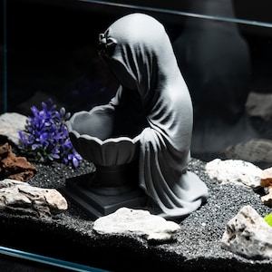 May include: A gray statue of a veiled figure holding a decorative bowl. The statue is placed in a fish tank with black gravel, rocks, and purple artificial plants. The figure is draped in flowing fabric.