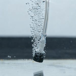 May include: Close-up of an aquarium air stone with a clear tube, releasing a stream of bubbles. The air stone is black and porous, designed to diffuse air into the water. The background is a gradient of light and dark gray.