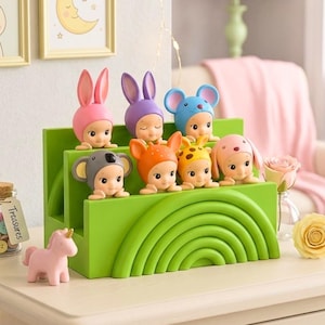 K&ouml;nnte beinhalten: Eine Sammlung kleiner Figuren, jede mit einem Hut im Tiermotiv, auf einem gr&uuml;nen, mehrstufigen St&auml;nder mit Regenbogenmuster ausgestellt. Eine rosa Einhornfigur und ein Glas mit bunten Kn&ouml;pfen sind ebenfalls vorhanden.