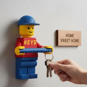 Puede incluir: Un llavero azul, rojo y amarillo estilo Lego montado en una pared, sosteniendo llaves. El llavero tiene la palabra "KEYS" en el frente. Un letrero de madera con "HOME SWEET HOME" está montado al lado. Una repisa debajo contiene una suculenta, carteras y gafas de sol.