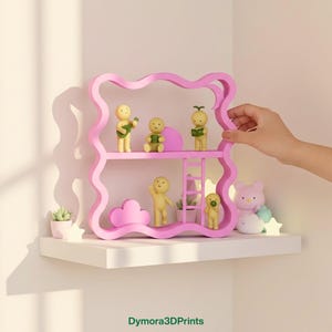 May include: A pink, wavy-edged shelf unit with two levels, holding small figurines and decorative items. The figurines are yellow and the shelf has a ladder. A hand is touching the shelf. The text "Dymora3DPrints" is at the bottom.