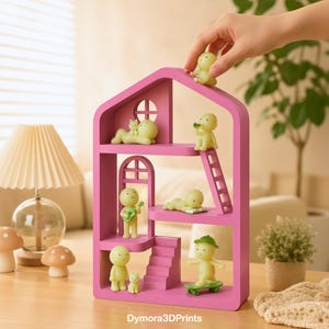 May include: A pink dollhouse-shaped shelf unit with multiple levels, featuring small, yellow figurines in various poses. Some figurines are playing instruments, skateboarding, or reading. The shelf has a window and a door. The text "Dymora3DPrints" is at the bottom.