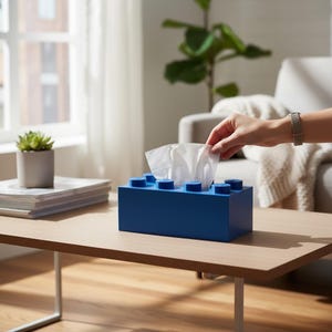 Può includere: Un dispenser di fazzoletti blu rettangolare progettato per assomigliare a un mattoncino. La scatola ha borchie blu rialzate sulla parte superiore. Una mano sta prendendo un fazzoletto. Su un tavolo di legno.
