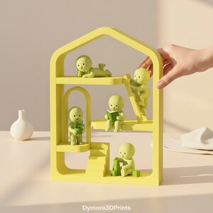 May include: A yellow, multi-tiered decorative shelf shaped like a house, featuring small green figurines in various poses. One figurine is reading a book, another is climbing a ladder, and others are interacting with blocks and plants. The shelf has an arched doorway.