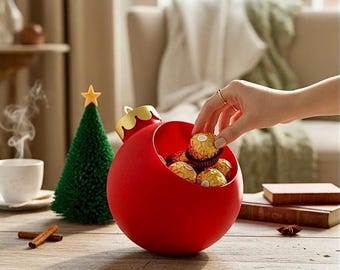 May include: A red, ornament-shaped container filled with wrapped chocolates. A small green Christmas tree with a gold star sits nearby. The container has a gold-colored top and is sitting on a wooden table. Cinnamon sticks and a cup of coffee are also on the table.