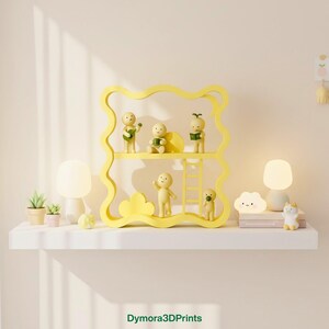 May include: A yellow, wavy-edged shelf unit displays small figurines, a ladder, and a cloud-shaped accent. Two small lamps and potted plants flank the shelf on a white surface. The text "Dymora3DPrints" is at the bottom.