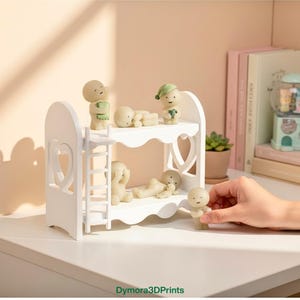 Op de afbeelding: Een wit stapelbed met hartvormige uitsparingen bevat verschillende kleine, beige beeldjes. Sommige beeldjes staan, andere liggen. E&eacute;n beeldje wordt door een hand vastgehouden. De beeldjes hebben eenvoudige kenmerken en staan in verschillende poses.