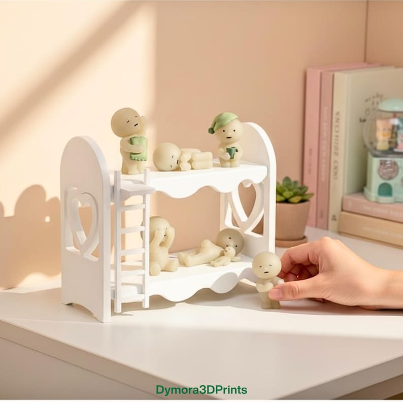 Op de afbeelding: Een wit stapelbed met hartvormige uitsparingen bevat verschillende kleine, beige beeldjes. Sommige beeldjes staan, andere liggen. E&eacute;n beeldje wordt door een hand vastgehouden. De beeldjes hebben eenvoudige kenmerken en staan in verschillende poses.