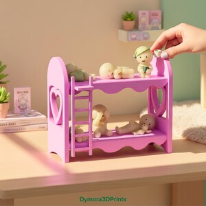 Op de afbeelding: Een roze miniatuur stapelbed met een hartvormige uitsparing, met daarin verschillende kleine beeldjes. De beeldjes bevinden zich in verschillende poses, sommige liggen, andere zitten. Een hand plaatst een beeldje op het bovenste bed. De tekst "Dymora3DPrints" staat onderaan.