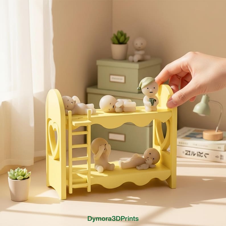 Op de afbeelding: Een gele stapelbed met hartvormige uitsparingen en een kleine ladder, bevolkt met miniatuurbeeldjes. De beeldjes bevinden zich in verschillende poses, sommige liggen, andere zitten. Een hand verstelt een van de beeldjes. De sc&egrave;ne speelt zich af op een lichtgekleurd oppervlak.