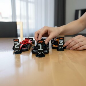 May include: A collection of miniature Formula 1 race cars in red, black, and orange. Each car has a corresponding number on a black and white checkered stand. The cars are arranged on a light brown surface.