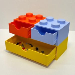 May include: A colorful storage unit designed to resemble a large building block. The unit features three pull-out drawers in orange, blue, and yellow, with a fourth drawer partially visible. The top of the unit has the classic block stud design.