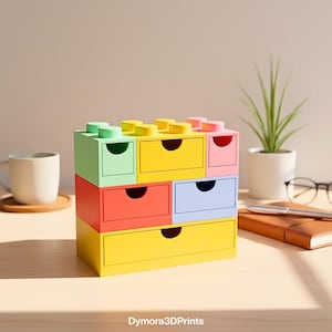 Op de afbeelding: Een veelkleurige opbergunit met meerdere lades, ontworpen om op een bouwsteen te lijken. De lades zijn in verschillende kleuren, waaronder groen, geel, rood, blauw en roze. De tekst "Dymora3DPrints" is zichtbaar onderaan.