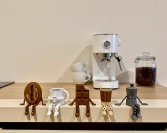 May include: A collection of whimsical coffee-themed figurines, including a coffee bean, a coffee cup, a chocolate bar, a coffee cup, and a stovetop espresso maker. A white espresso machine, coffee cups, and a jar of coffee beans are in the background.