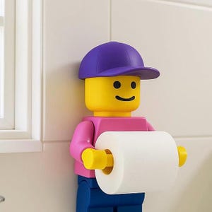 May include: A novelty toilet roll holder designed as a Lego figure. The figure has a yellow head with a smiling face, a purple cap, a pink shirt, blue trousers, and yellow arms. It holds a roll of white toilet paper.
