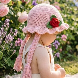 May include: A light pink crochet hat with a red rose and green leaves, featuring two long, braided tassels. The hat is worn by a child in a beige romper, sitting on a blanket in a garden setting.