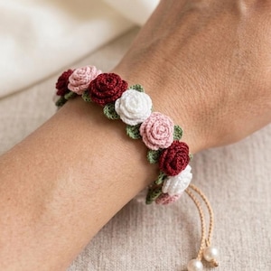 Puede incluir: Pulsera de ganchillo con diseño floral. Presenta rosas de ganchillo en rojo, rosa y blanco, con hojas verdes. Es ajustable con un cordón beige y perlas. Un accesorio encantador.