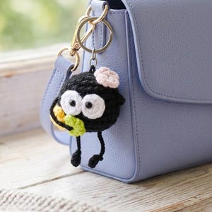 May include: A handmade black crochet keychain in the shape of a cartoon character with large white eyes, a pink flower, and small legs. The keychain is attached to a light blue purse with a gold-colored clasp.