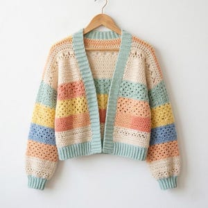 Könnte beinhalten: Ein farbenfroher, offener Strickcardigan mit Streifenmuster. Der Pullover hat Pastelltöne in Blau, Gelb, Pfirsich und Creme mit hellblauen Verzierungen. Der Cardigan wird auf einem Holzbügel vor weißem Hintergrund präsentiert.