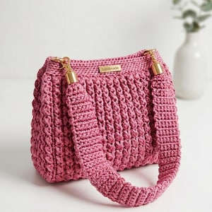 May include: A dusty rose crochet handbag featuring a textured, ribbed stitch pattern. The bag includes a thick, matching crochet shoulder strap attached with gold-toned metal hardware. A small rectangular metal tag labeled handmade is centered.