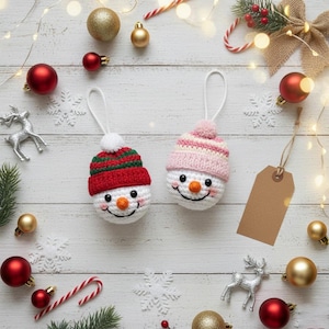 May include: Two handmade snowman ornaments with smiling faces, wearing red and green or pink and white knit hats. The ornaments are surrounded by Christmas decorations, including ornaments, snowflakes, and candy canes, on a white wooden surface.