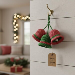 May include: Three crocheted Christmas bells in red and green hang from a brass hook. Each bell has a white bead inside. A small brown tag with the words "Merry & Bright" is attached to the bells. The bells are hanging on a white wooden wall.