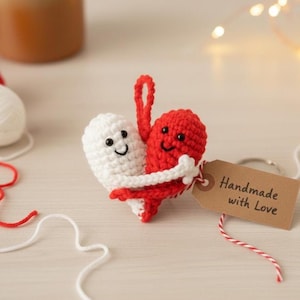 May include: A handmade crochet heart in red and white, hugging. The white heart has a smiling face, and the red heart has a smiling face. A small tag reads "Handmade with Love".