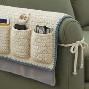 May include: Cream-colored, crocheted sofa armrest organizer with three pockets. The organizer has a blue trim and two wooden buttons. A gray fabric panel hangs down. The organizer is tied to the sofa arm with a cream-colored rope.