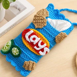 May include: A blue crocheted tissue holder shaped like a potato chip bag, featuring a yellow and red oval patch with the white text "lays" in the center. The design includes small crocheted brown chips and green lime slices as decorative accents.