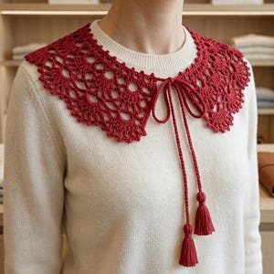 Puede incluir: Un cuello de ganchillo rojo con un borde festoneado y un cierre de lazo con borlas. El cuello se lleva sobre un suéter color crema.