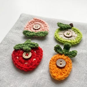 May include: Four handmade crochet fruit appliques: a peach, apple, strawberry, and carrot. Each fruit is a different colour, with green leaves and a wooden button centre. The appliques are on a grey fabric background.