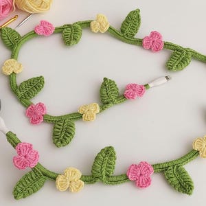May include: A crocheted cable protector in the shape of a vine, adorned with green leaves and pink and yellow flowers. The cable protector is designed to wrap around a charging cable, adding a decorative touch. The background includes yarn and a black object.