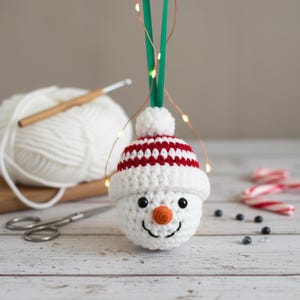 May include: A crocheted snowman ornament with a red and white striped hat, black eyes, and an orange nose. The snowman is hanging from a green ribbon. A ball of white yarn, a crochet hook, and scissors are in the background.