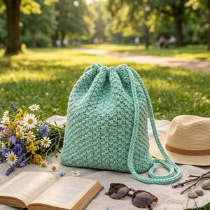 May include: A mint green crochet drawstring bag with a braided strap sits on a blanket with a straw hat, sunglasses, and an open book. A bouquet of wildflowers is next to the book. The scene is set in a park.