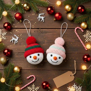 Peut inclure: Deux décorations de bonhomme de neige faites à la main avec des chapeaux rouges et roses, des visages souriants et des pompons. Les ornements sont entourés de décorations de Noël, dont des boules, des branches de pin et des lumières, sur une surface en bois.