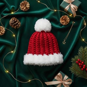 May include: A red and white knitted Christmas hat with a white pom-pom. The hat has a white fluffy trim. The hat is on a green satin surface, surrounded by gold pine cones, a star necklace, and wrapped gifts.
