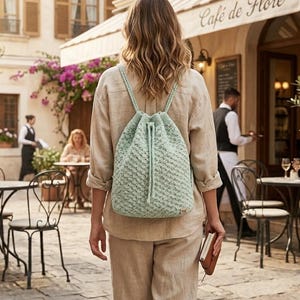 May include: A light blue knitted drawstring backpack with a textured pattern. The backpack has braided straps and a drawstring closure. The person is walking in front of a cafe with the text "Cafe de Flore".