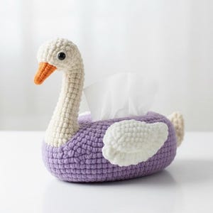 May include: A handmade crochet tissue box cover shaped like a swan. The swan has a white head and neck, an orange beak, and a purple body with white wing accents. White tissues are visible from the opening.