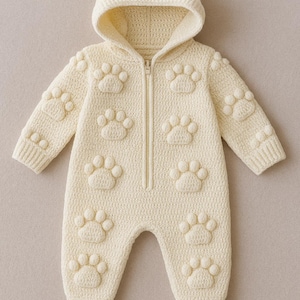 May include: Cream-coloured baby jumpsuit with a hood and a full-length zip. The knitted jumpsuit features raised paw prints across the front, sleeves, and legs. The hooded onesie is made of a soft, textured material.
