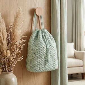 May include: A pale green drawstring bag with a textured finish hangs from a wooden peg on a wooden wall. The bag has a drawstring closure and a woven appearance. The background includes a vase of dried flowers and a curtain.