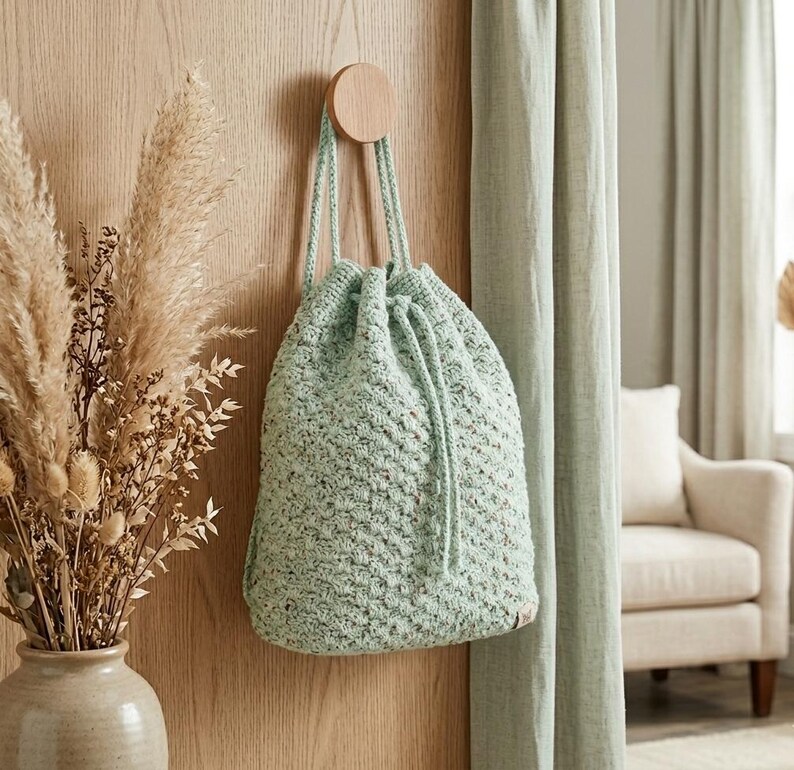 May include: A pale green drawstring bag with a textured finish hangs from a wooden peg on a wooden wall. The bag has a drawstring closure and a woven appearance. The background includes a vase of dried flowers and a curtain.