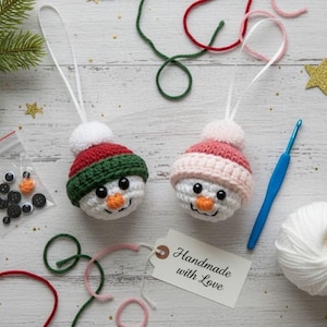 May include: Two handmade crochet snowman ornaments with red, green, and pink hats, white pom-poms, and orange carrot noses. The scene includes craft supplies like yarn, a crochet hook, and a tag that says "Handmade with Love."