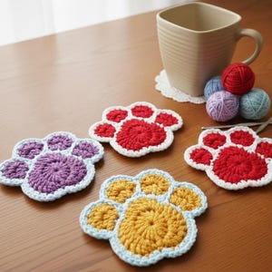 May include: Four handmade crochet paw print coasters in red, purple, and yellow with white and blue trim. A beige mug, yarn balls, and crochet hooks are also on the wooden table. The coasters are approximately 4 inches in diameter.