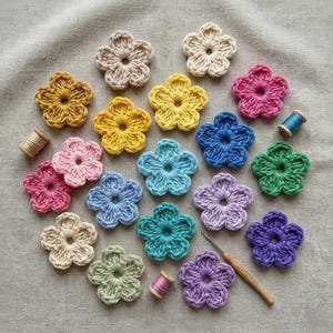 Può includere: Una collezione di applicazioni floreali all'uncinetto colorate in varie tonalità, tra cui giallo, rosa, blu e viola. I fiori fatti a mano sono disposti su una superficie di tessuto neutro, con un uncinetto e rocchetti di filo visibili.