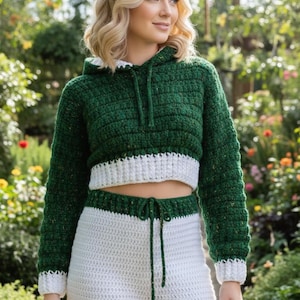 May include: A green and white crochet hoodie and matching trousers set. The cropped hoodie has a drawstring and white trim. The trousers have a drawstring waist and white trim. The outfit is made of a textured yarn.