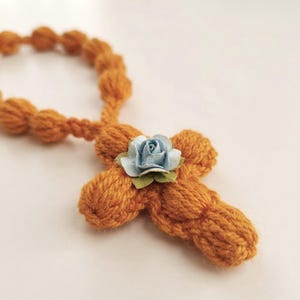 May include: A crocheted rosary made of mustard yellow yarn with a series of rounded beads and a central cross shape. A small, light blue artificial rose with green fabric leaves is attached to the center of the cross, adding a floral accent.
