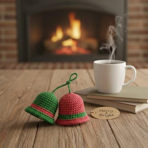 May include: Two crocheted bell ornaments, one green and one red, with green and red stripes, sit on a wooden surface next to a white mug of coffee and a tag that says "Merry & Bright". A fireplace is in the background.