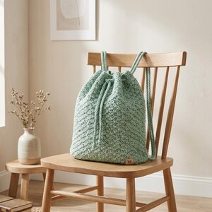 May include: A light green, hand-crocheted drawstring backpack with matching drawstrings and straps. The bag is sitting on a wooden chair. The bag has a textured appearance and a small, brown leather tag.