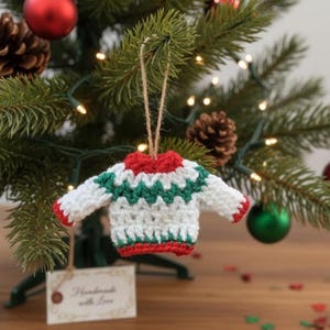Può includere: Un ornamento natalizio fatto a mano a forma di maglione all'uncinetto. Il maglione bianco ha motivi a zig-zag verdi e rifiniture rosse. È appeso a una corda su un albero con decorazioni e pigne.