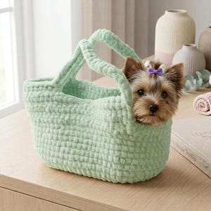 May include: A light green, hand-crocheted tote bag with two handles. A small Yorkshire Terrier dog with a purple bow is peeking out of the bag. The bag is sitting on a wooden surface.